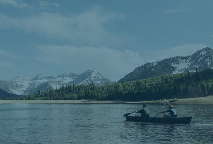 Guys in a canoe on a pretty lake. Nice trees. Mountains with snow in the background.