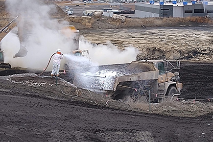 A person in a hazmat suit blowing chemicals from a hose onto the soil in a dump truck. A backhoe and steam are in the background.