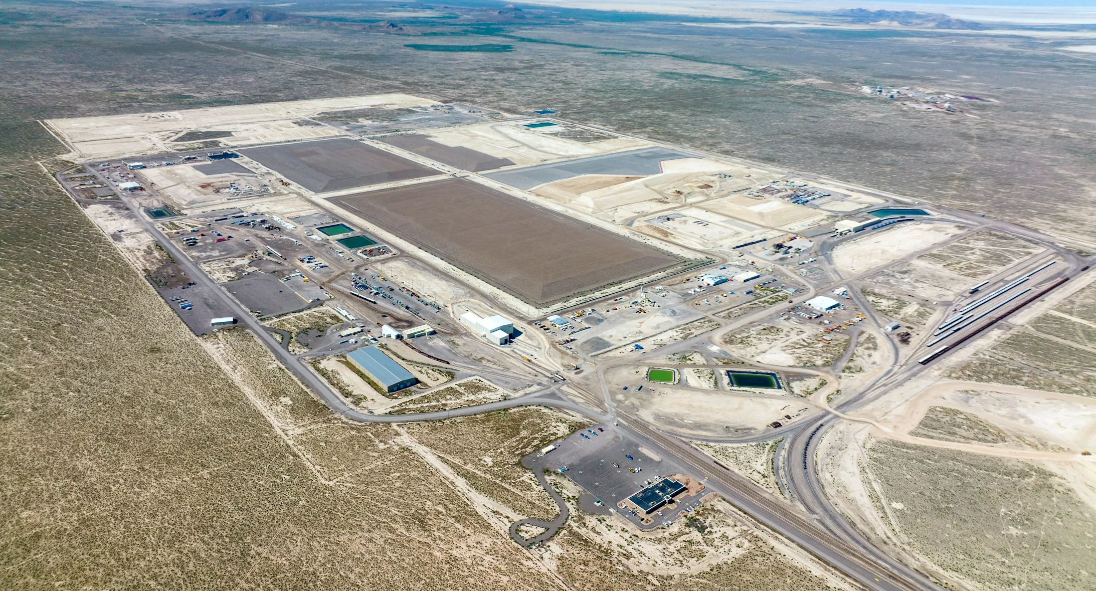 Aerial wide-angle view of the EnergySolutions Clive facility, a large-scale low-level radioactive waste disposal site located in a remote, flat desert landscape in Tooele County, Utah. The image shows a sprawling industrial complex organized into large, rectangular disposal cells (embankments) with different levels of capping and fill. The facility is heavily integrated with transportation infrastructure, featuring an extensive network of internal dirt and paved roads, a dedicated rail spur with multiple tracks for offloading waste shipments, and various support buildings, specialized handling facilities, and evaporation ponds. The surrounding environment is arid and uninhabited, stretching to distant mountains under a clear sky, emphasizing the site's isolation and vast operational footprint.